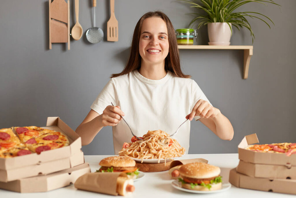 Horizontal shot of delighted beautiful hungry woman with brown hair wearing white casual T-shirt sitting at table in kitchen, eating pasta, looking at camera with toothy smile. - Foto, Bild
