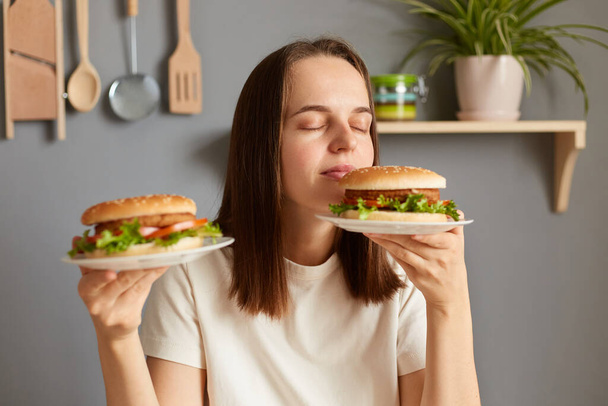 Portrait of brown haired Caucasian woman holding two delicious fresh hamburgers, smelling aromat of fast food with closed eyes and satisfied facial expression. - Foto, Imagem