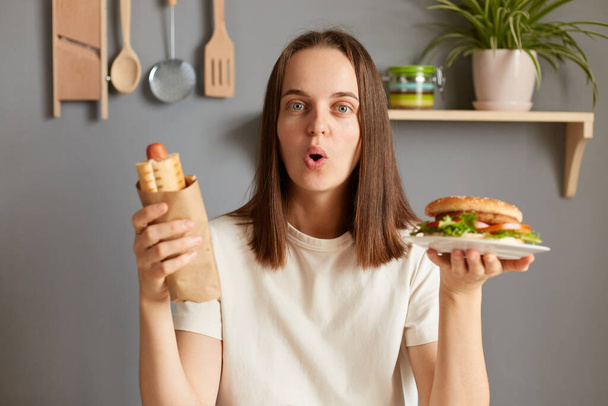 Surprised amazed woman with dark hair eating delicious burger and hot dog, breaking diet and likes fast food, wearing white T-shirt, looking at camera, saying wow, looks astonished. - Fotó, kép