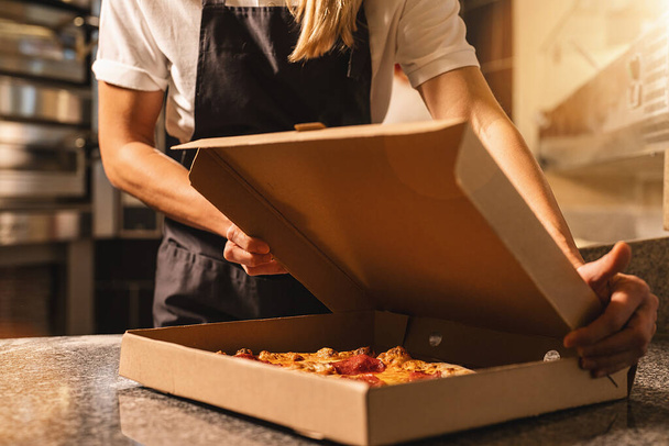 female pizza baker packing a fresh pizza from the stone oven in a box for delivery at italian pizzeria - Foto, afbeelding