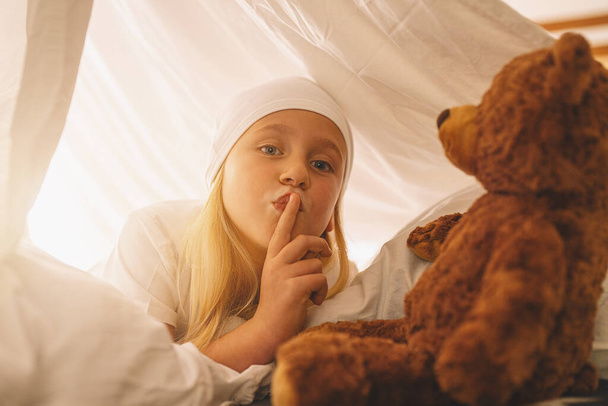 Γλυκό κοριτσάκι με αρκουδάκι κράτα το δάχτυλο στα χείλη δείχνοντας σιωπή για να κρατήσεις ένα μυστικό ενώ ξαπλώνει στο κρεβάτι της σε μια σκηνή με ποτό.. - Φωτογραφία, εικόνα