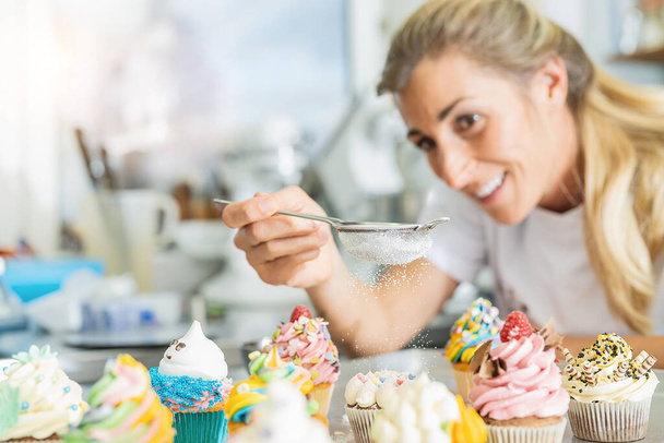 happy women in pastry bakery as confectioner sprinkles powdered sugar in a colander as topping on a colorful muffin - Foto, Imagem