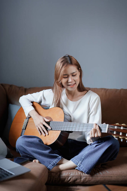 Портрет молодой азиатки, играющей на гитаре на диване расслабляющий стресс в отпуске. - Фото, изображение