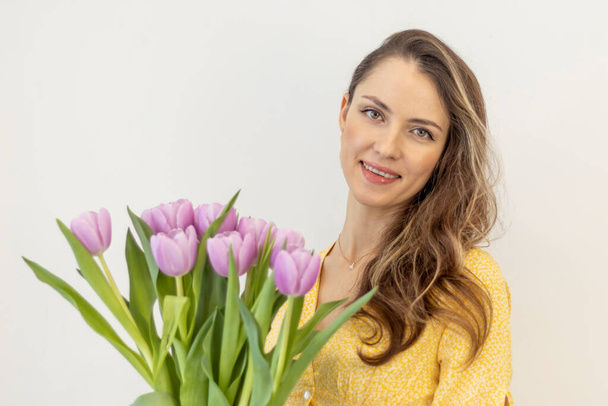 Portret van een mooie lachende vrouw in het geel met een boeket lila tulpen op een witte achtergrond. - Foto, afbeelding