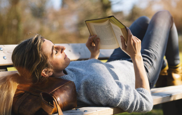 Читающая женщина лежит в парке на своей сумке на скамейке. студент, читающий концепцию книги - Фото, изображение