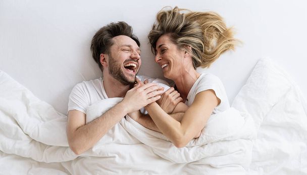 Vista superior de pareja joven y cariñosa acostada en la cama riendo y sonriendo con manta. Se miran con confianza en el amor. - Foto, Imagen