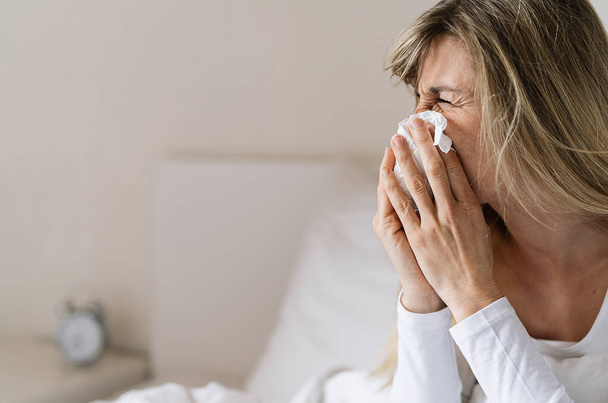 Choroba kobiety siedzącej na łóżku pod kocem, zamarzającej, cieknącej z nosa, przeziębionej kichającej w chusteczce, chorej dziewczynie z objawami grypy kaszącej w domu, obrazem grypy - Zdjęcie, obraz