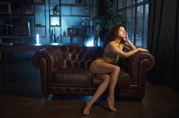 Barefoot sensual woman with eyes closed wearing sexy lingerie with yellow laces sitting on leather sofa - Foto, imagen