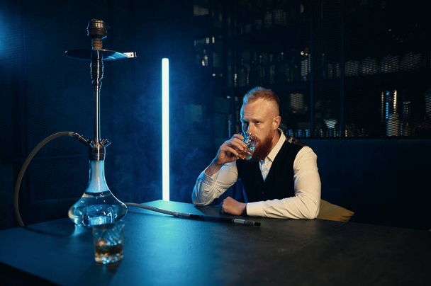 Stylowy facet w garniturze pijący whisky ze szkła w barze Hookah. Portret pewnego siebie bogatego biznesmena - Zdjęcie, obraz
