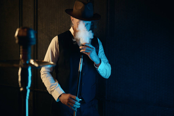 Front view of resting man smoking hookah at home workplace. Retro guy in hat drinking relax in dark room with neon lighting - Foto, Bild