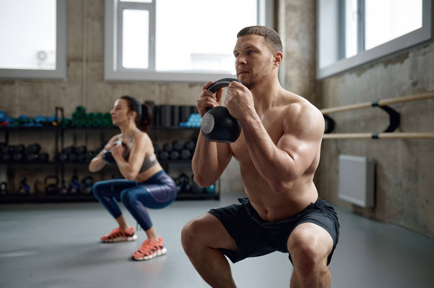 Man and woman with good physique holding heavy kettle bell for swing cross training hard core workout in gym. Personal class with professional sport coach - Foto, Imagen