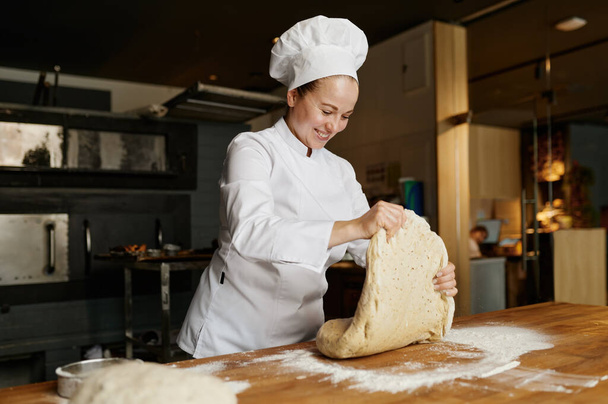 Female baker kneading or forming dough while working at bakehouse. Fresh pizza, pie, bread or sweet pastry preparation process - Foto, Bild