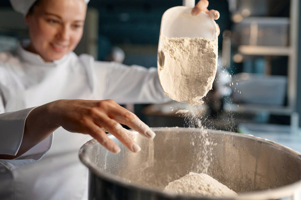 Profesjonalny cukiernik noszący biały mundur wkładający mąkę do dużej metalowej miski stojącej przy stole w cukiernictwie. Widok z bliska, ostrość selektywna - Zdjęcie, obraz
