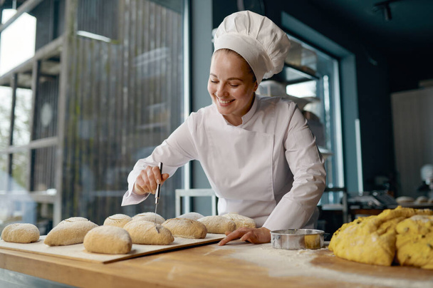 Woman baker wearing uniform engaged in making of traditional yeast buns with cheese - Фото, изображение