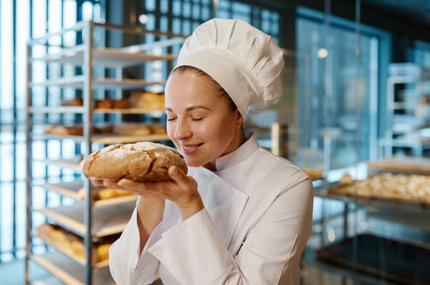 Молодая пекарь нюхает аппетитный свежий хлеб на фоне современной коммерческой пекарни. Производство выпечки - Фото, изображение