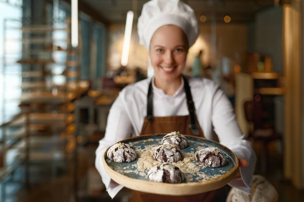 Onnellinen naiskondiittori esittelee tuoreita keksejä leipomokeittiössä. Onnellinen makeinen näyttää herkullisia tuoreita makeita suklaaleivoksia tarjottimella - Valokuva, kuva