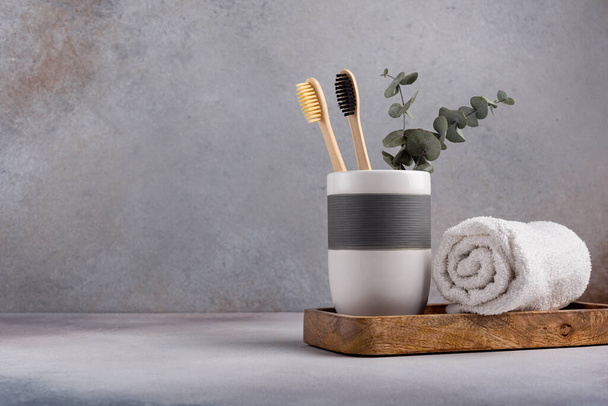 Eco friendly bathroom concept, sustainable lifestyle. Couple of bamboo toothbrush in a glass with white towel and eucalyptus on wooden tray - Фото, зображення