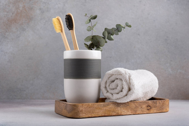 Eco friendly bathroom concept, sustainable lifestyle. Couple of bamboo toothbrush in a glass with white towel and eucalyptus on wooden tray - Zdjęcie, obraz