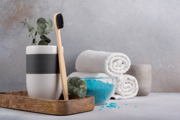 Eco friendly bathroom concept, sustainable lifestyle. Bamboo toothbrush in a glass with white towels, eucalyptus on wooden tray and blue sea salt - Zdjęcie, obraz