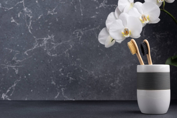 Eco friendly bathroom concept. Couple of bamboo toothbrush in a glass with white orchid flowers on dark grey background with copy space - Foto, immagini
