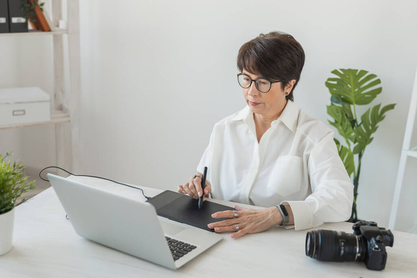 Keski-ikäinen nainen graafinen suunnittelija tai valokuvaaja, joka käyttää digitaalista graafista tablettia työskennellessään nykyaikaisessa toimistossa, ammattimainen naisten korjaaja, joka istuu nykyaikaisessa työtilassa, geneerinen muotoilu tabletti ja - Valokuva, kuva