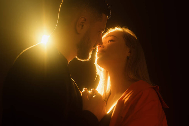 Oldalnézet fiatal szakállas férfi és szőke hajú nő fogja egymás kezét, és csók óvatosan a sötétben ellen izzó fény - Fotó, kép