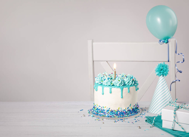 Blue and white birthday party with a buttercream drip cake, sprinkles, bright gold candle, hat and balloons on a light grey background. Trendy. Copy space.  - Zdjęcie, obraz