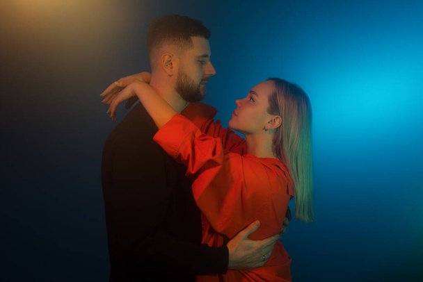 Vista lateral del joven hombre y la mujer en ropa casual tocándose mientras están de pie en habitación oscura con iluminación de neón azul - Foto, imagen