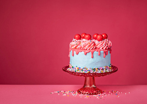 Bubble Goma de mascar Cumpleaños Drip Cake en un soporte rojo con chispas de colores, glaseado de crema de mantequilla y bolas de caramelo de cereza roja sobre un vibrante fondo rosa. Por encima de la diversión. Copiar espacio. - Foto, imagen