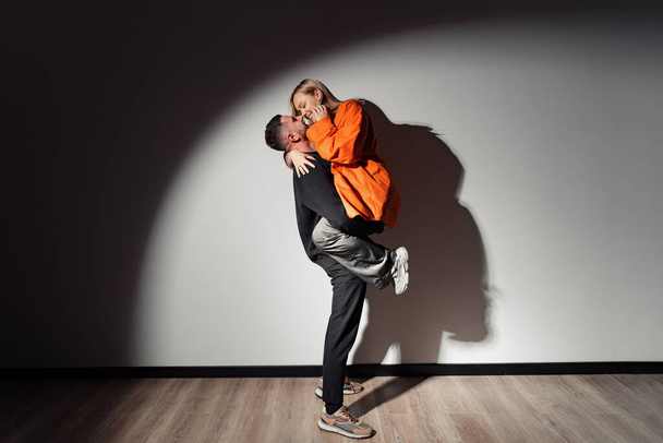 Vista lateral de cuerpo completo del hombre joven levantando y abrazando a la mujer mientras toca las narices en la habitación oscura iluminada por la lámpara - Foto, Imagen