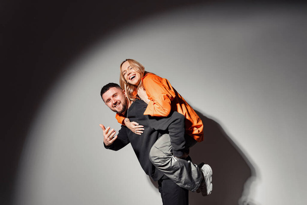 Side view of cheerful man in black clothes giving piggyback ride to excited girlfriend wearing orange jacket and raising hand in dark room illuminated by round lamp - Photo, Image