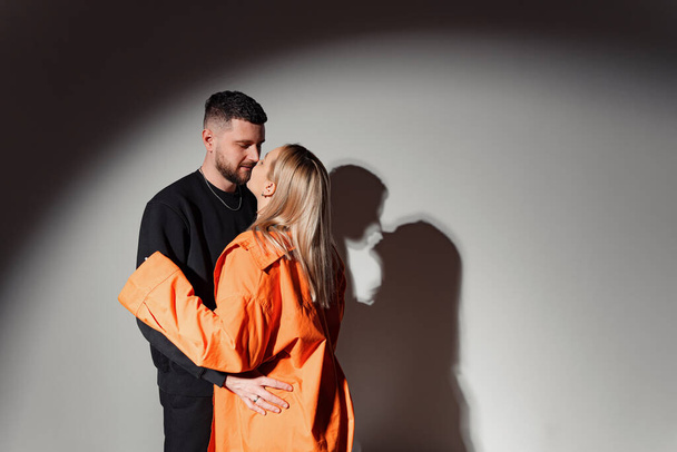 Vista laterale del giovane che abbraccia e bacia la fidanzata contro il muro con l'ombra dalla luce incandescente in uno studio buio - Foto, immagini
