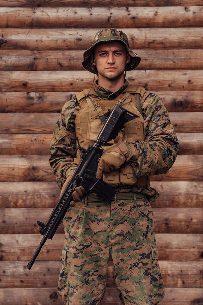A soldier in uniform with a rifle in his hand is standing in front of a wooden wall. A soldier guards the forest base from the enemy. - Фото, изображение