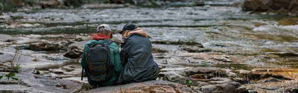 Rear, couple of hikers sitting on bank of rocky river, resting when hiking in mountains. Back view. - Foto, imagen
