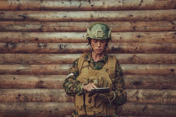 Woman soldier using tablet computer against old wooden wall in camp. - Фото, изображение