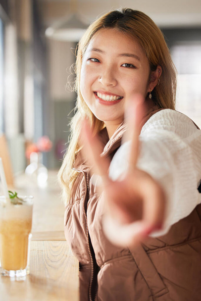 Peace, hand gesture and portrait with an asian woman in a coffee shop, drinking a beverage or refreshment. Face, emoji and cafe with an attractive young female enjoying a smoothie or juice drink. - Фото, изображение