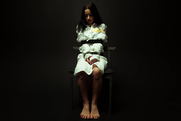 Una mujer mentalmente inestable y enferma con una camisa de fuerza sentada en una silla en un hospital psiquiátrico sobre un fondo negro aislado. Concepto de trastornos mentales. Preson loco. - Foto, Imagen