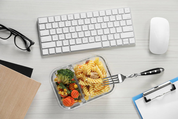 Container with tasty food, keyboard, mouse, notebooks and glasses on white wooden table, flat lay. Business lunch - Valokuva, kuva
