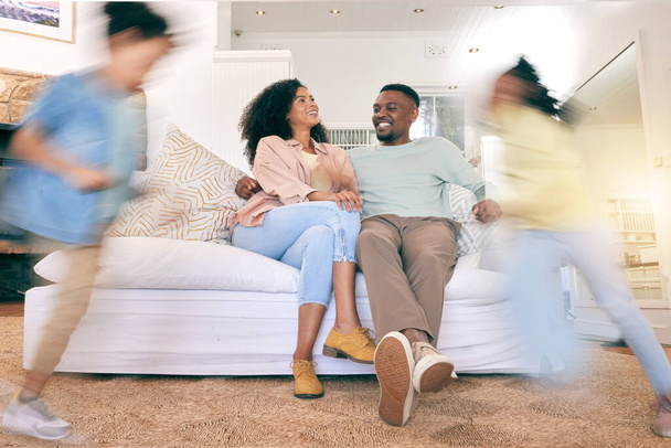 Children running, happy home and couple on a sofa, talking and relax while bonding in a living room. Blur, kids and content parents on a couch, smile and enjoying weekend indoors with playing and fun. - 写真・画像