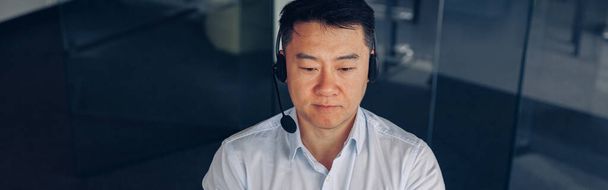 Aasialainen liikemies kuulokkeissa työskentelee tietokoneella modernissa coworking. Laadukas kuva - Valokuva, kuva