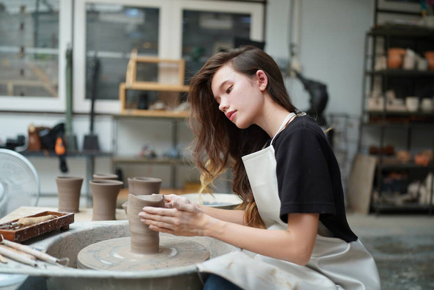 Close-up de artesã bonita concentrada em avental sentado na roda de cerâmica e usando ferramenta artesanal ao moldar vaso de barro molhado - Foto, Imagem