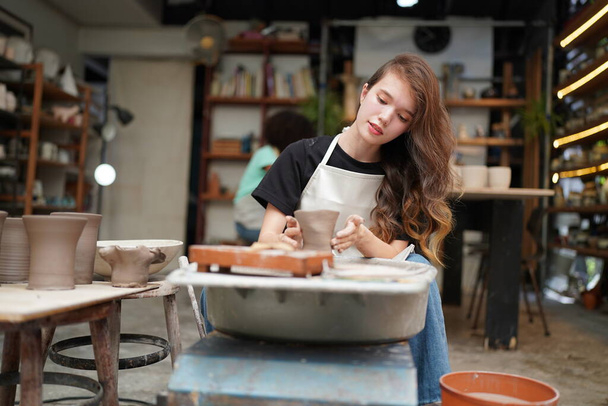 Close-up of concentrated beautiful craftswoman in apron sitting at pottery wheel and using craft tool while shaping wet clay vessel - 写真・画像