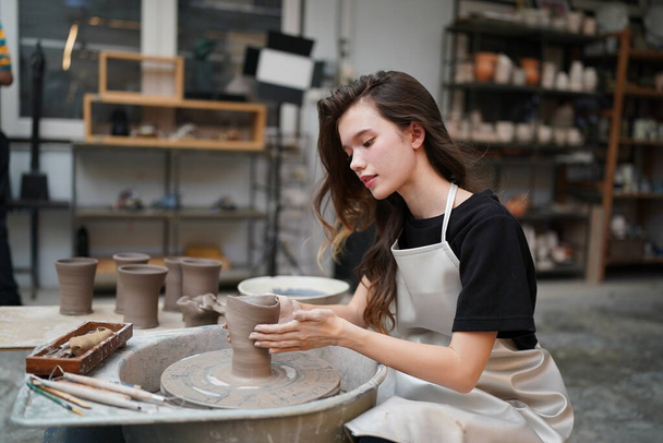 Close-up of concentrated beautiful craftswoman in apron sitting at pottery wheel and using craft tool while shaping wet clay vessel - Photo, Image