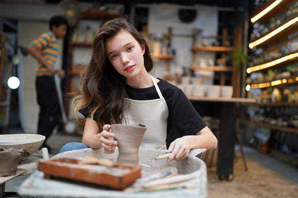 Close-up of concentrated beautiful craftswoman in apron sitting at pottery wheel and using craft tool while shaping wet clay vessel - Фото, изображение