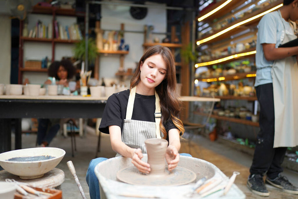 Close-up of concentrated beautiful craftswoman in apron sitting at pottery wheel and using craft tool while shaping wet clay vessel - Фото, изображение