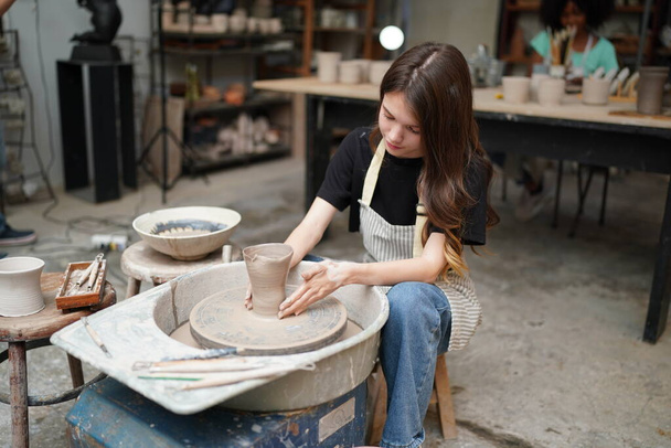 Primer plano de la hermosa artesana concentrada en delantal sentada al volante de cerámica y usando una herramienta artesanal mientras moldea un recipiente de arcilla húmeda - Foto, imagen