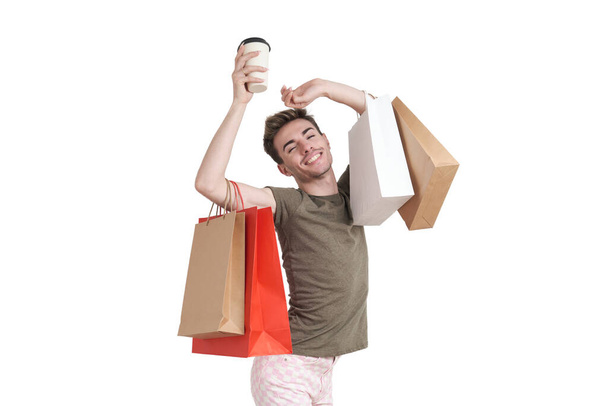 Beyaz arka planda izole edilmiş alışveriş torbaları ve kahve taşıyan genç beyaz bir adam dans ediyor.. - Fotoğraf, Görsel