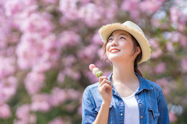 春の桜まつりの期間中、桜の木の公園を歩いている間に甘い花見団子デザートを持っているアジアの女性 - 写真・画像