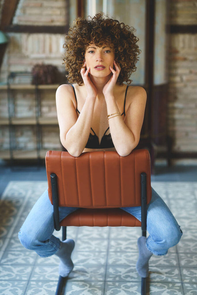 Full body of sensual young female, with afro hairstyle in black lingerie and blue jeans witting on leather chair and touching face while looking at camera against brick wall - Foto, Bild