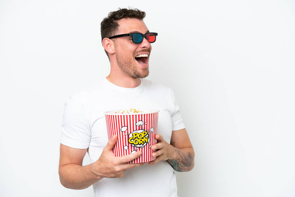 Young caucasian handsome man isolated on white background with 3d glasses and holding a big bucket of popcorns - Foto, Imagen